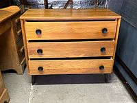 Front view of wooden 3-drawer chest of drawers showing three drawers with black handles and short tapered legs.