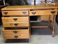 Front angled view of the wooden desk showing all drawers, one drawer open at bottom left, metal handles, and turned legs on right side.