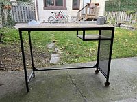 Front view of the metal frame desk with wooden top, showing the storage shelf and castor wheels.