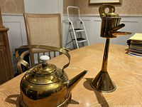 Photo showing two vintage metal items on a wooden table: a brass teapot with white knob lid and a tall brass oil lamp with wick holder and wide base.