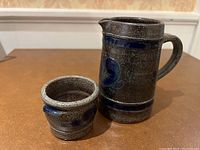 Photo showing small dark speckled earthenware carafe with handle and spout beside matching small cup, both with blue decorative accents