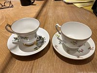 Two bone china teacups and saucers placed side by side on a wooden table showing floral designs and gold trim