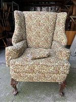 Front view of the floral upholstered vintage wingback chair showing the winged armrests, cushion and carved cabriole wooden legs.