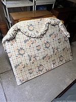 Floral upholstered headboard with plaid and flower pattern, decorative ruffle, curved top edge, and plywood backing visible at the bottom.