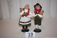 Full view of both figurines showing girl and boy in traditional German clothing, each holding a gingerbread man, standing on green bases with a number 19 card visible.