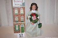 Full view of Mrs. Fezziwig figurine next to a small display card and box showing her name and a green door. The figurine wears a light green striped dress and holds a wreath with bows.
