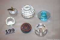 All six paperweights arranged together, showing variety of shapes and colors including clear faceted ball, blue mushroom, clear bubble with signature, millefiori, apple shape and clear with bubbles.
