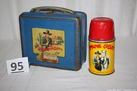 Photo showing the blue metal Hopalong Cassidy lunch box with handle and clasp alongside the matching yellow Aladdin thermos with red lid and Hopalong Cassidy printed on the front.