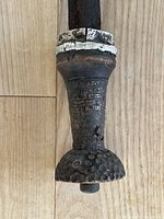 Close-up of the sword handle showing a turned wooden grip decorated with carved geometric patterns and copper wire wrapping, exhibiting wear and some damage.