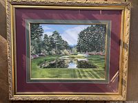 Framed print showing the golf course with detailed surroundings, gold ornated frame, maroon matting, and signature visible