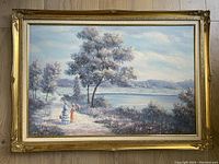 Full view of the vintage oil painting in ornate gold frame, showing the entire landscape scene with two women walking by a lake path under trees.