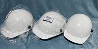 Photo of three white hard hats, two wrapped in plastic and one without wrapping showing exterior shell shape and style.