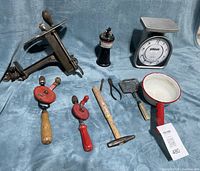 Photo of the vintage lot including antique meat cutter, Pelouze kitchen scale, vintage torch, hand drills, tin pot, hammer, soap cage and pliers set on fabric background