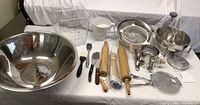Wide view of baking and cooking utensils laid out including large stainless bowl, food mill, strainers, rolling pins, tortilla press, knives and spatulas