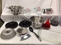Full lot overview showing all kitchen and baking items including mixing bowls, Pyrex measuring cups, colander, bundt cake pans, segmented baking pans, kitchen scale, funnels, cooking spoon, and glass baking dish.