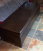 Front angled view of the dark brown faux leather storage bench placed on tiled floor next to couch
