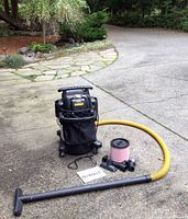 Full view of Dewalt shop vacuum with long yellow hose, black wand, multiple attachments, pink filter, and manual on driveway surface.