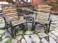 Front view of two connected patio rockers with attached central table, showing weathered wood and metal frames with moss.
