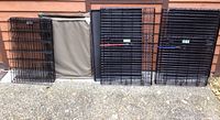 Two large folded black metal wire dog crates, a folded black metal 8-panel dog fence, a beige dog bed, and folded black mat resting against an exterior house wall.