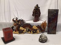 Photo showing entire lot including carved dragon, samurai figurine, large dark orange candle on plate, small ceramic statue and tall rectangular Raku ceramic wall art.