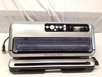 Front and top view of FoodSaver vacuum sealer, silver and black unit with buttons and sealing area visible.