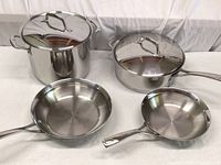 Full set of stainless steel cookware including one large stock pot with lid, a large pot with lid, and two skillets arranged on a cloth surface.