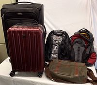 All items shown together on table with white backdrop: large black Samsonite soft luggage, maroon Delsey hard carry-on luggage, two black and gray backpacks, and olive Fossil messenger bag.
