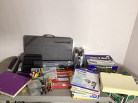Photo showing assorted office supplies including pens, markers, file folder labels, Scotch tape, staplers, 3-hole punch, canned air, and various paper products with a black plastic lap desk