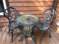 Full view of the wrought iron bistro table and two chairs showing black ornate scrollwork design and surface wear with green patina.