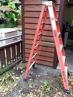 Side view of the 6-foot orange Werner step ladder showing fiberglass frame and aluminum steps, standing on gravel outside a wooden structure.