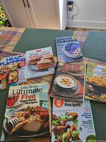 Overview of all seven cookbooks and magazines arranged on a table, showing colorful covers and titles related to meal planning, diabetes, slow cooking, and Weight Watchers.