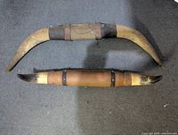 Two sets of Texas Longhorn cow horns laid on a carpeted surface. The larger set measures approximately 32 inches, the smaller set approximately 24 inches. Both sets show leather and metal band accents.