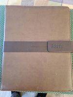 Front view of the brown leather Roots portfolio showing the magnetic close tab with embossed Roots branding.