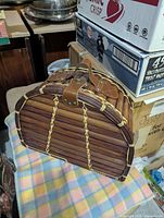 Closed view of brown wooden slatted picnic basket with leather-like handle and strap, sitting on a multi-colored plaid blanket.