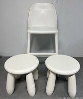 White children's highchair standing behind two white round stools with three legs each, against a gray background.