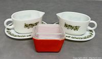 Front view of two Pyrex milk glass gravy boats with matching plates displaying the green Spring Blossom pattern and a small red square glass container with a clear lid in front.