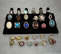 Top-down view showing entire collection of 31 assorted rings in multiple rows on display stand and flat surface. Various designs, colors and stones visible.