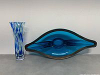 Side view of two items on a shelf: left is a tall multicolor swirl glass vase, right is a blue and amber centerpiece bowl with a leaf shape and glossy finish