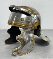 Side view of a silver-colored gladiator helmet with brass detailing and cheek guards, showing the visor brim and riveted brass studs.