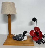 Wide shot of the wooden table lamp with beige shade, carved wood duck, and the vintage red spotlight lamp all together on a white surface.