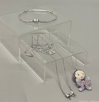 Swarovski necklace with butterfly doll face pendant and silver ball chain, alongside Pandora bracelet on a clear acrylic riser.