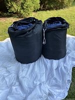 Two rolled Coleman sleeping bags in black carry bags placed on a white blanket outdoors with grass in the background.