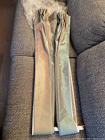 Two narrow, long drapery panels folded and laid on gray upholstery with taupe and green/gray stripes visible