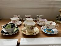 Front view of all nine teacups and saucers arranged in two rows on wooden surface, showing varied floral designs and rim shapes