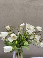 Photo of the artificial bouquet displaying white tulip flowers and green fronds in a clear vase against a plain wall background.