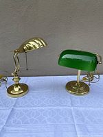 Photo of both lamps side by side on a white tablecloth showing the green glass shade bankers lamp on right and brass tone clamshell lamp on left.