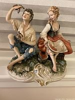 Full front view of the Capodimonte porcelain figurine depicting a young man holding grapes and a young woman holding watermelon sitting on a detailed base.