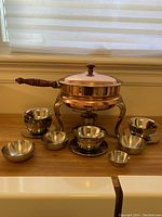 Full view of copper chafing dish with wooden handle and seven stainless steel bowls arranged around it on a wooden surface.