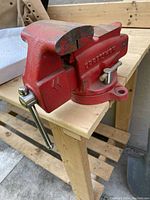 Side angle of red painted heavy metal Craftsman bench vice showing the fixed jaw, movable jaw, and clamp handle on a light wooden workbench.