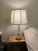 Full view of the lamp with fabric shade on, showing the lamp lit and placed on a wooden side table next to a bed.